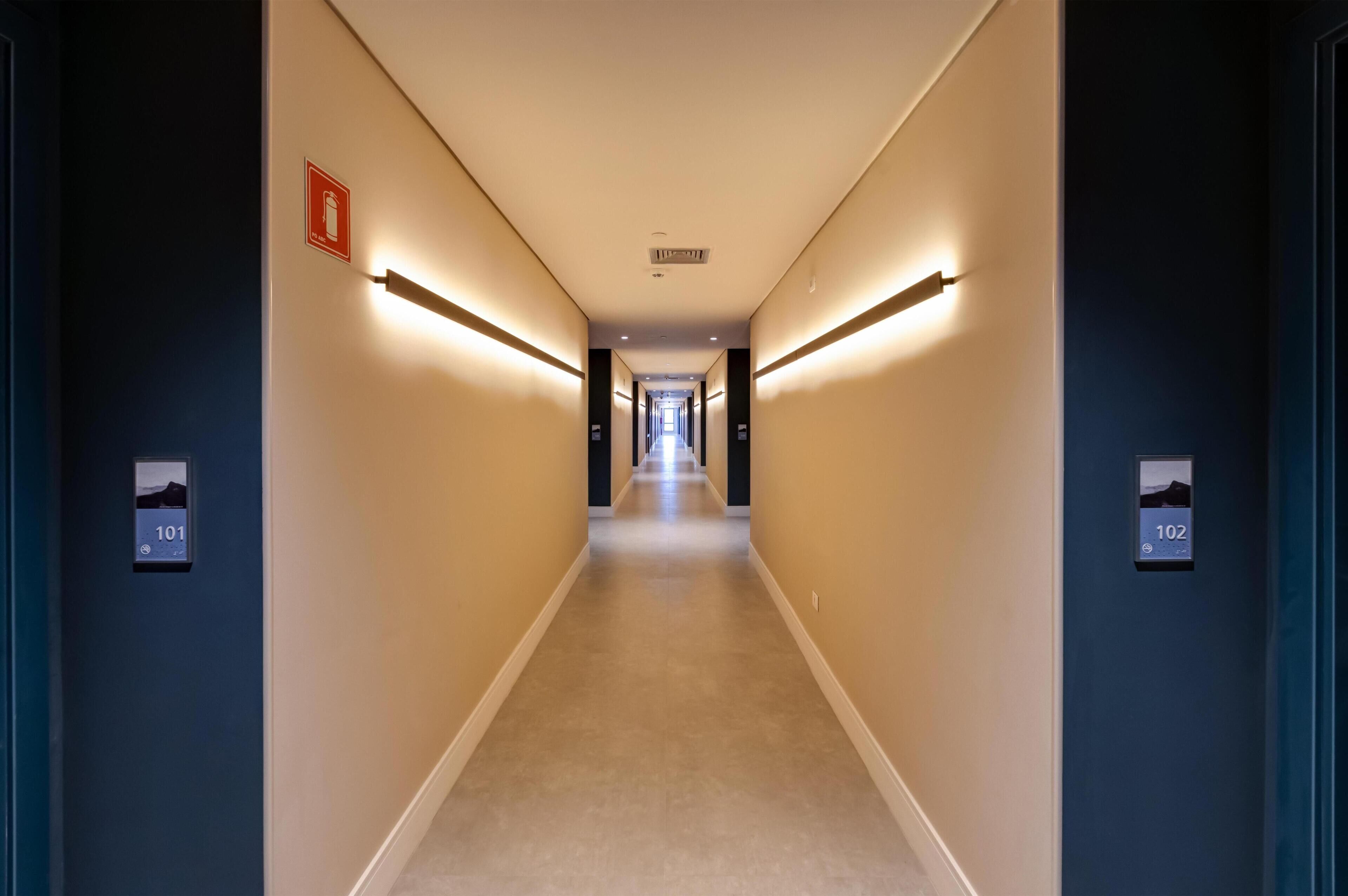 A hotel hallway with a beige carpet, dark walls, and illuminated handrails. There are signs indicating room numbers 101 and 102.