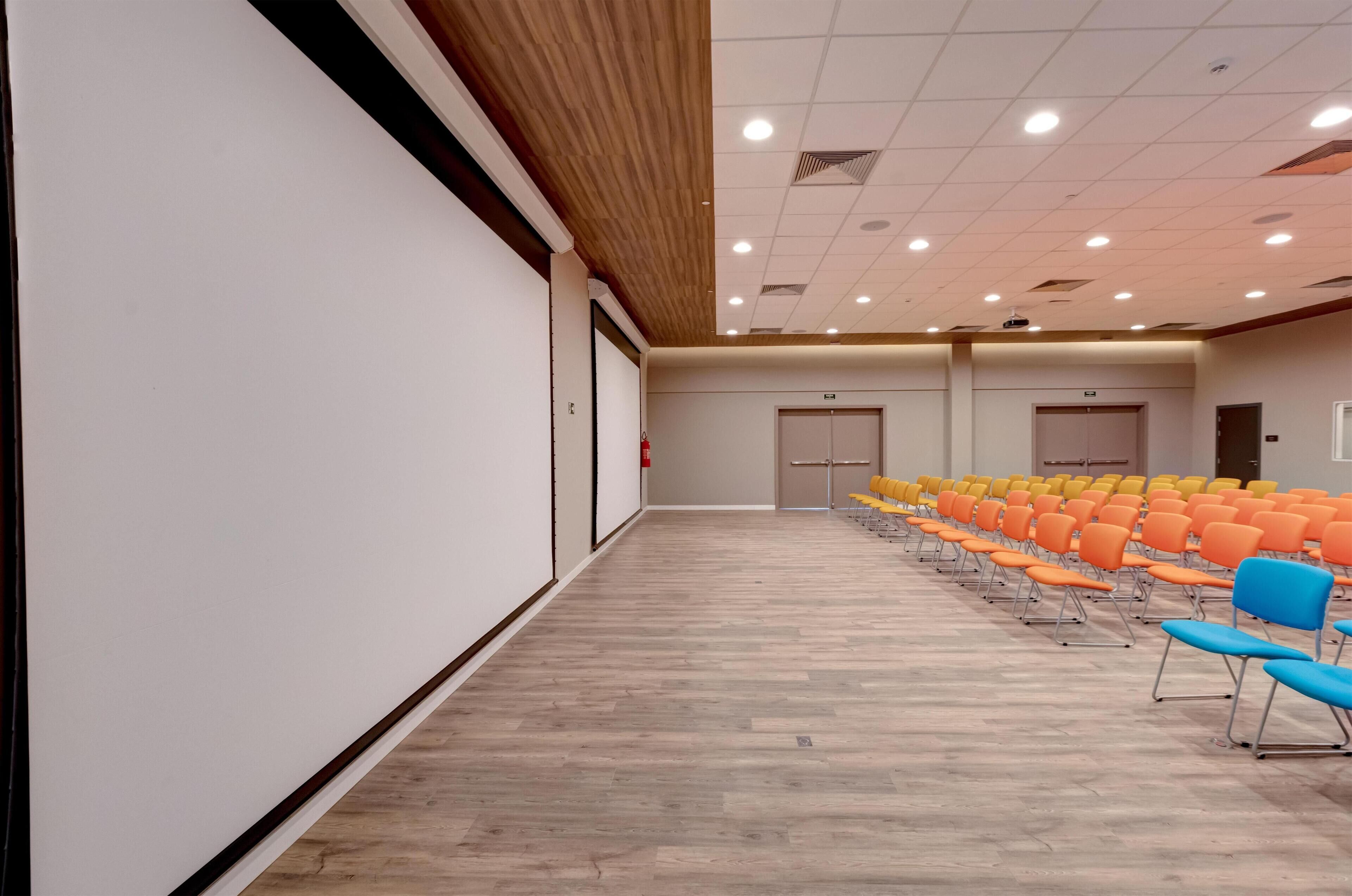 A conference room with rows of orange and blue chairs, wooden flooring, and a whiteboard on the left wall.