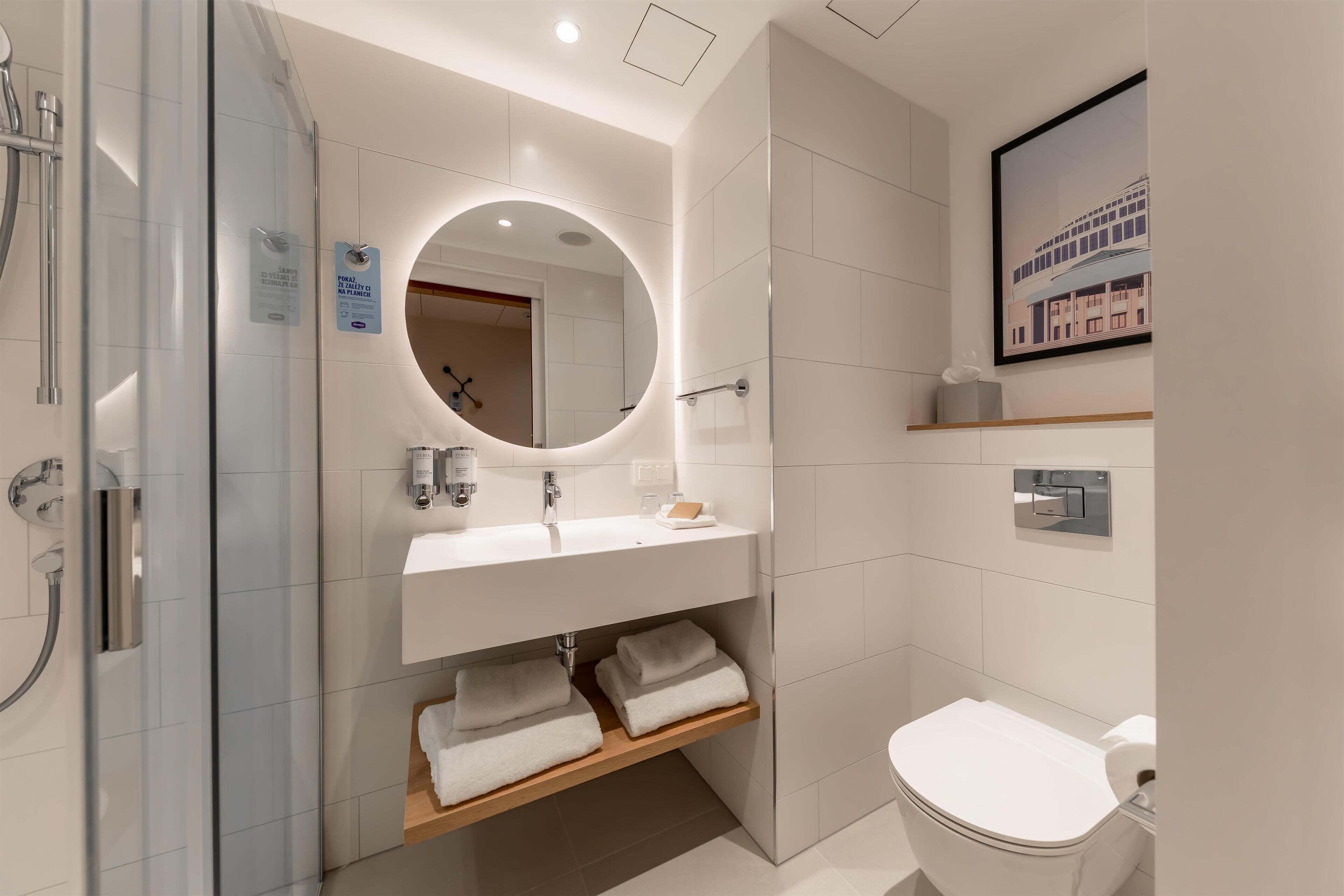 A modern bathroom with a glass shower enclosure, a round mirror, a white sink, and a toilet.