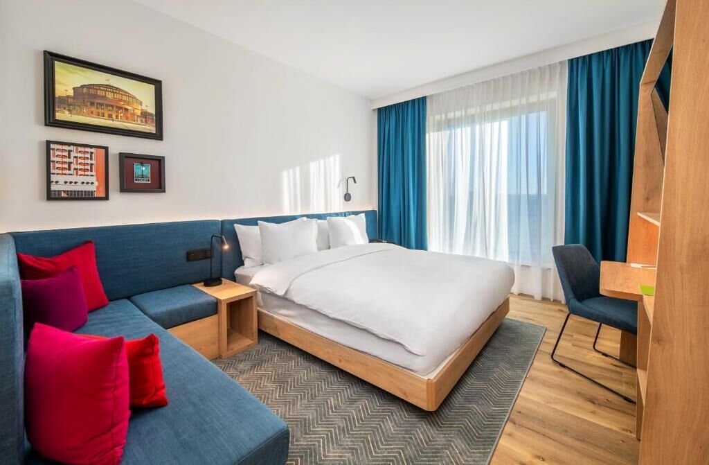 Une chambre d’hôtel avec un lit, un canapé, un bureau et une chaise. Des tableaux encadrés ornent le mur.