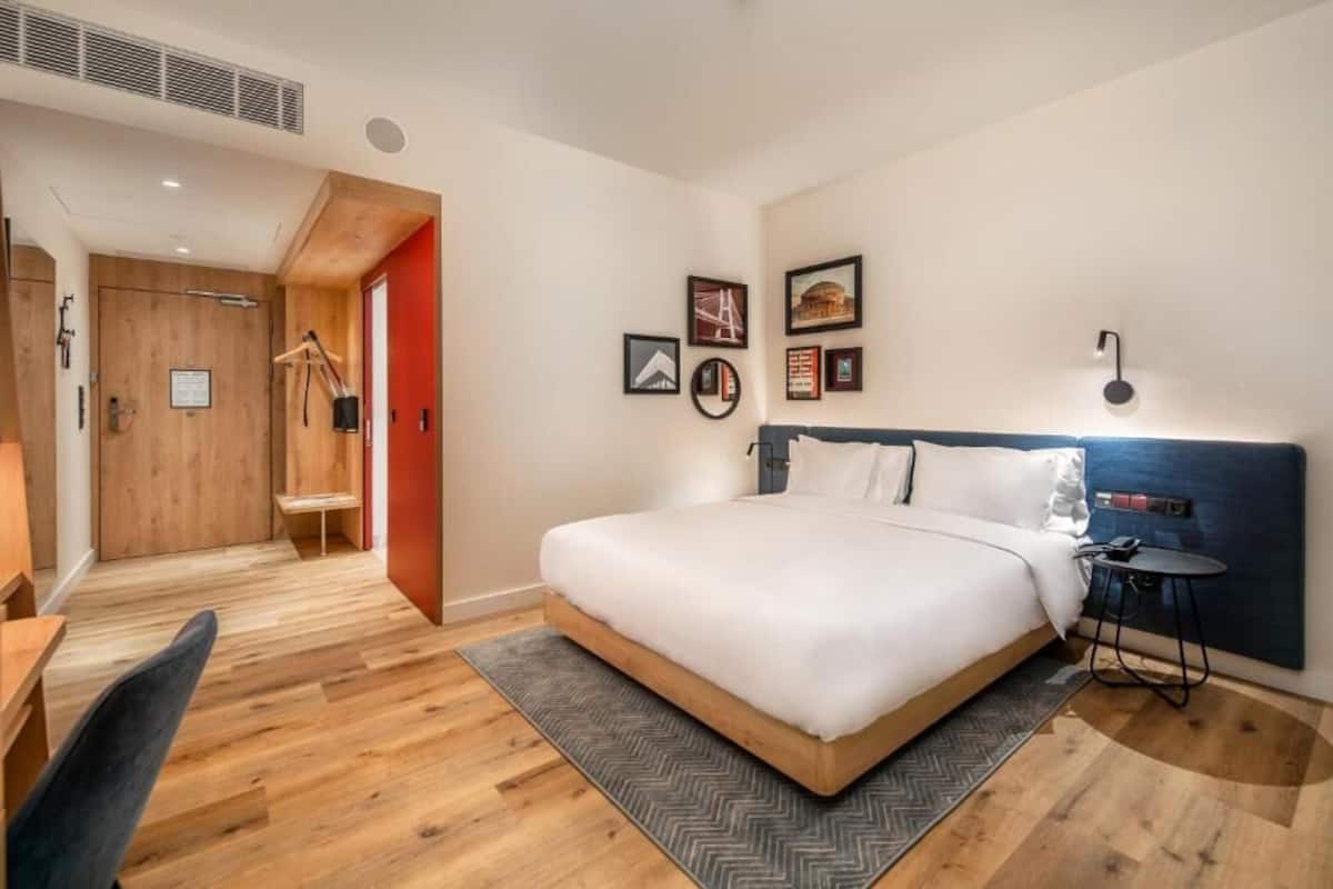 Quarto de hotel moderno com uma cama grande, uma escrivaninha, uma cadeira, uma mesinha de apoio e um guarda-roupa.