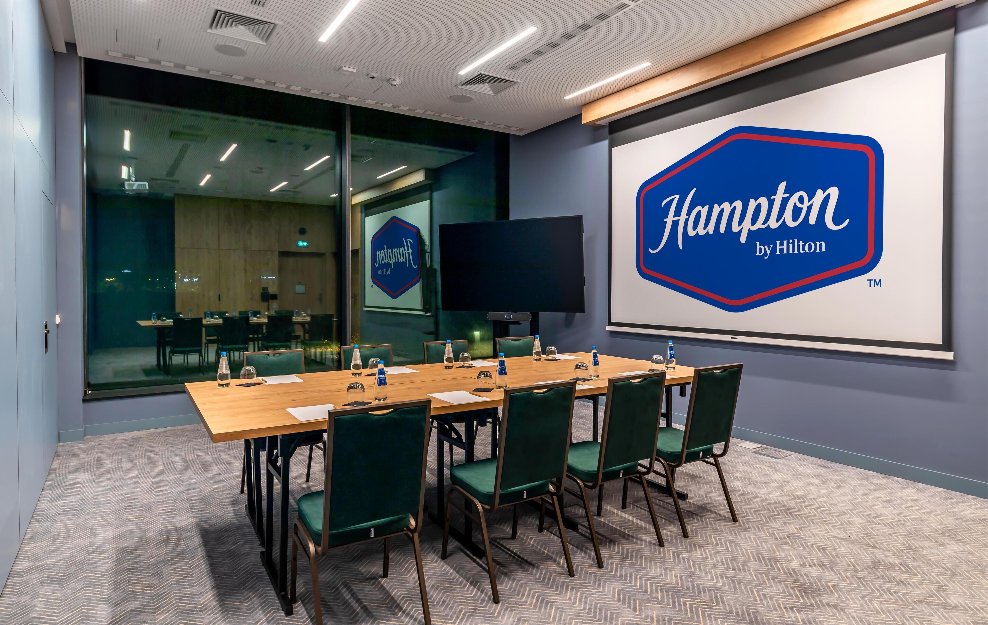 Un salón de conferencias con una pantalla grande que muestra el logotipo de 'Hampton by Hilton', una mesa de madera con sillas y un muro de vidrio con vista a la ciudad.