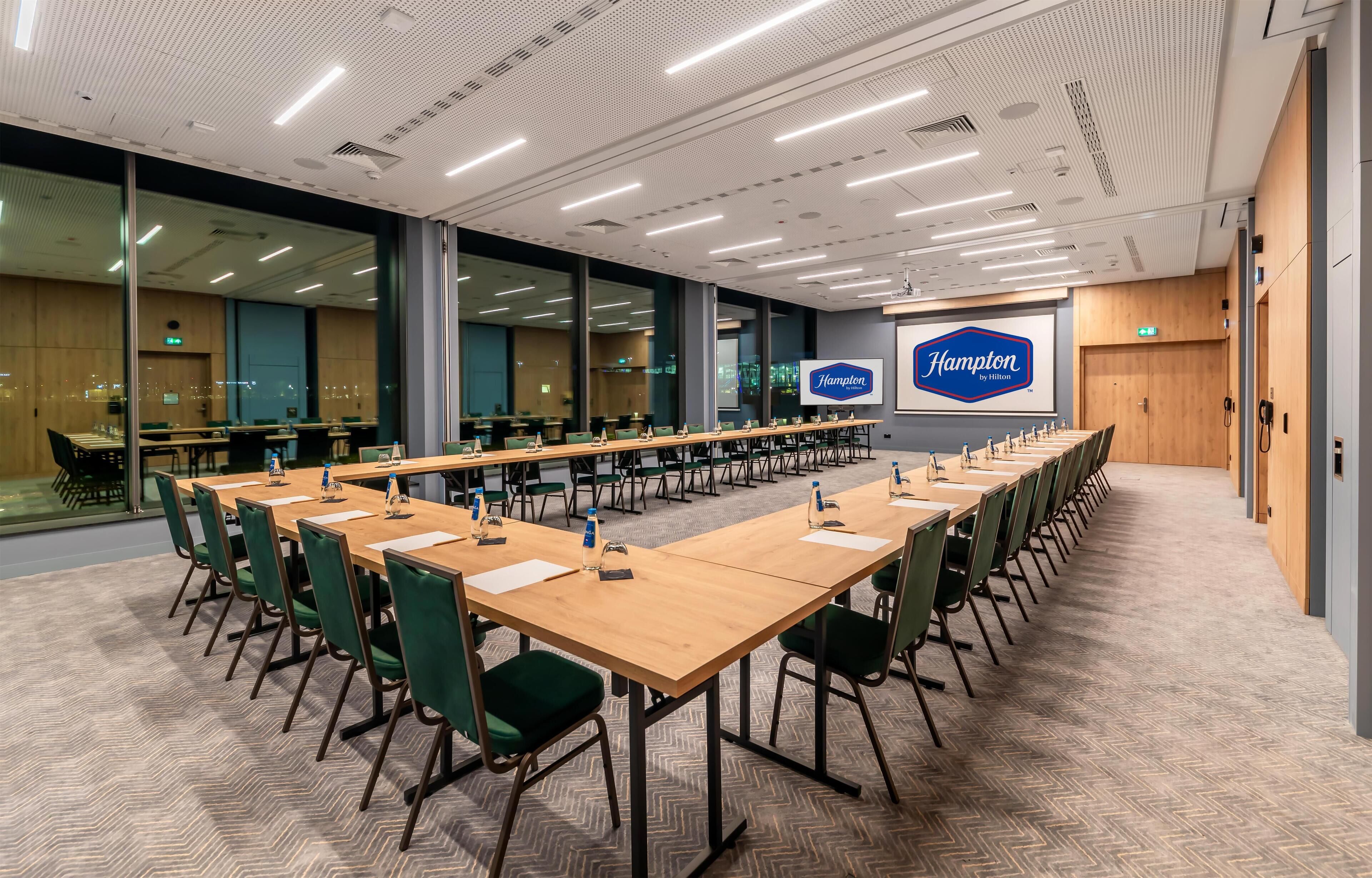 A conference room with long tables, green chairs, and a large screen displaying the Hampton by Hilton logo.