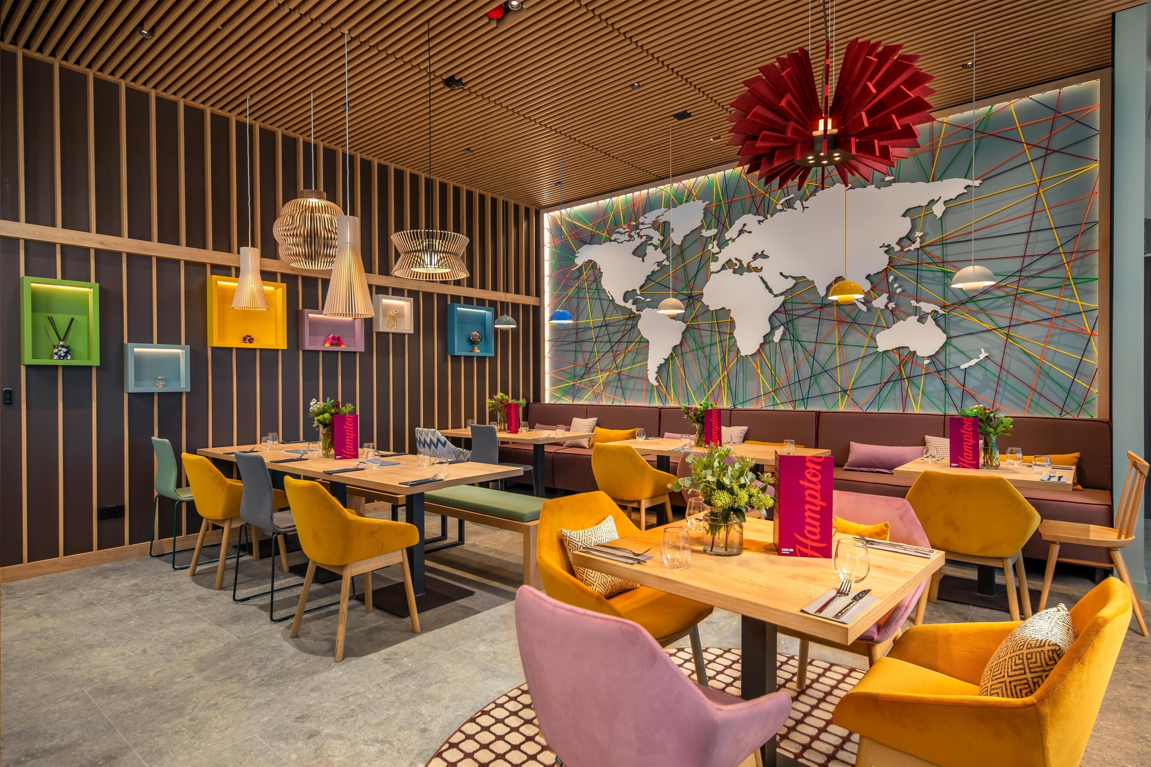 A modern restaurant with wooden panel walls, colorful chairs, and a large world map mural.