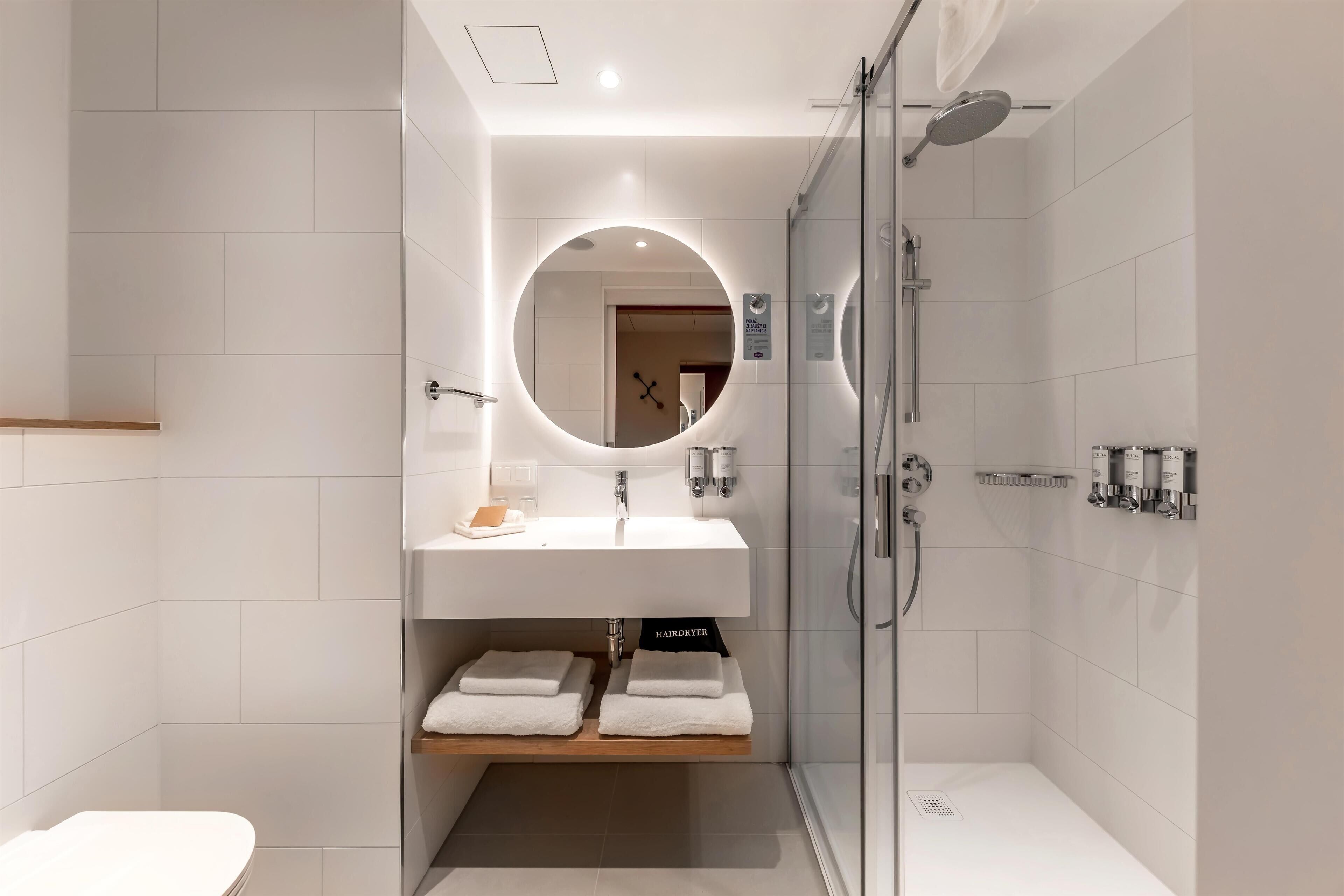 Une salle de bain moderne avec une douche à parois de verre, un miroir rond et un lavabo blanc surmonté d’une étagère en bois.