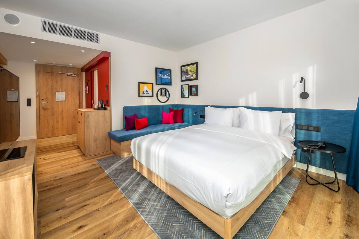 Ett modernt hotellrum med en stor säng, en sittgrupp med röda kuddar, trägolv och en blå accentvägg.