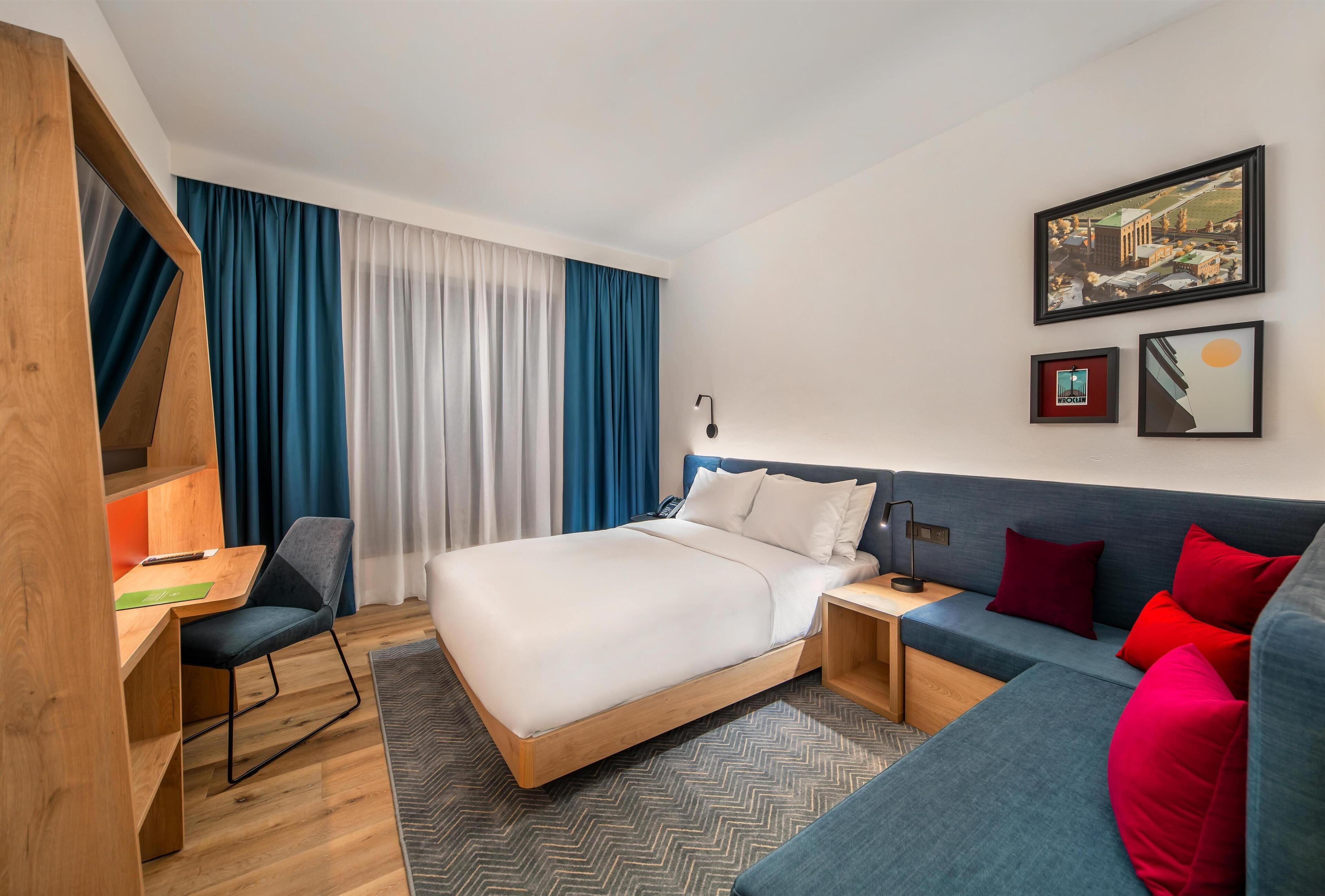 A modern hotel room with a bed, a desk, a chair, a sofa, and a large window with curtains.