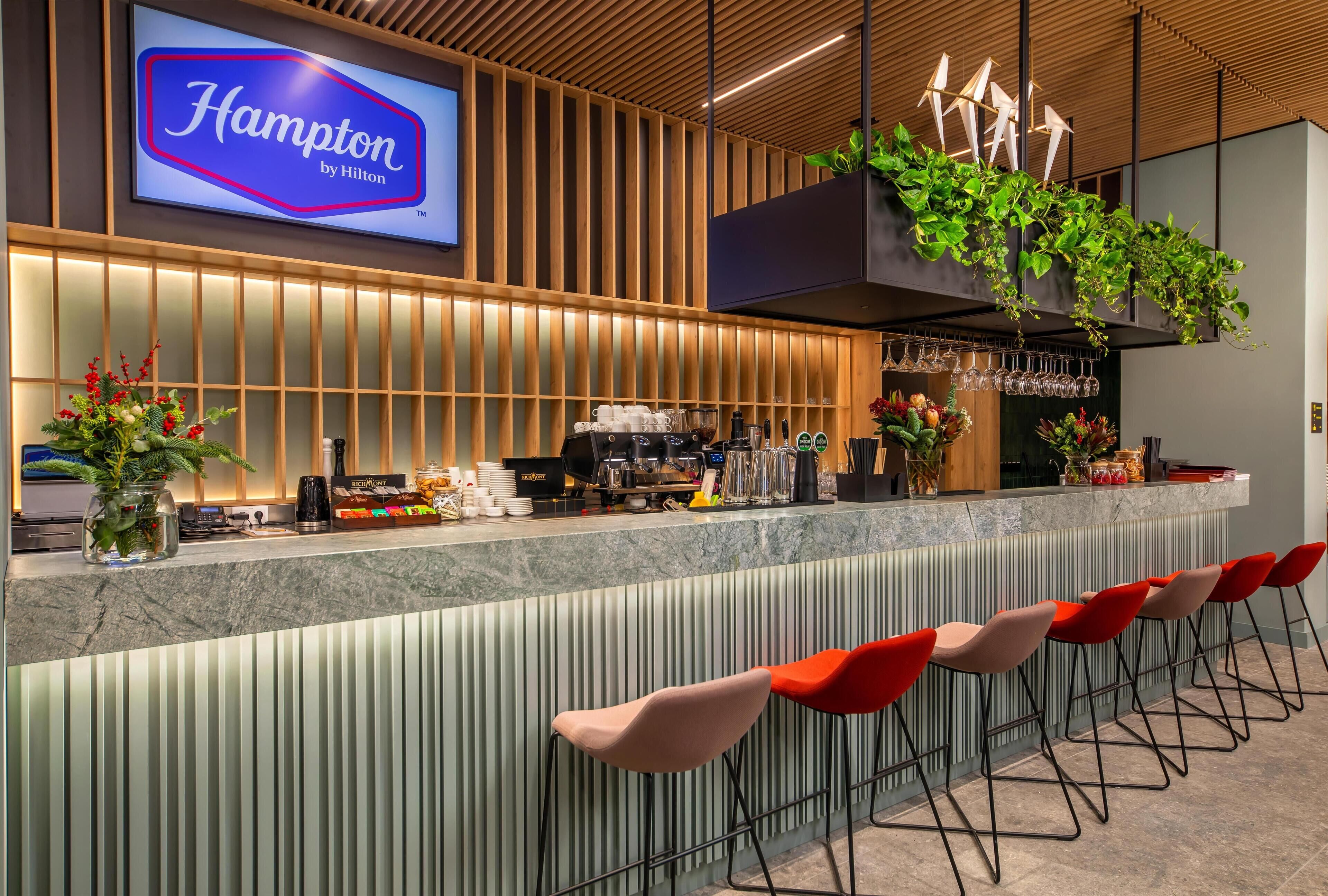 A modern bar area with a sign reading "Hampton by Hilton," bar stools, a coffee machine, and decorative plants.