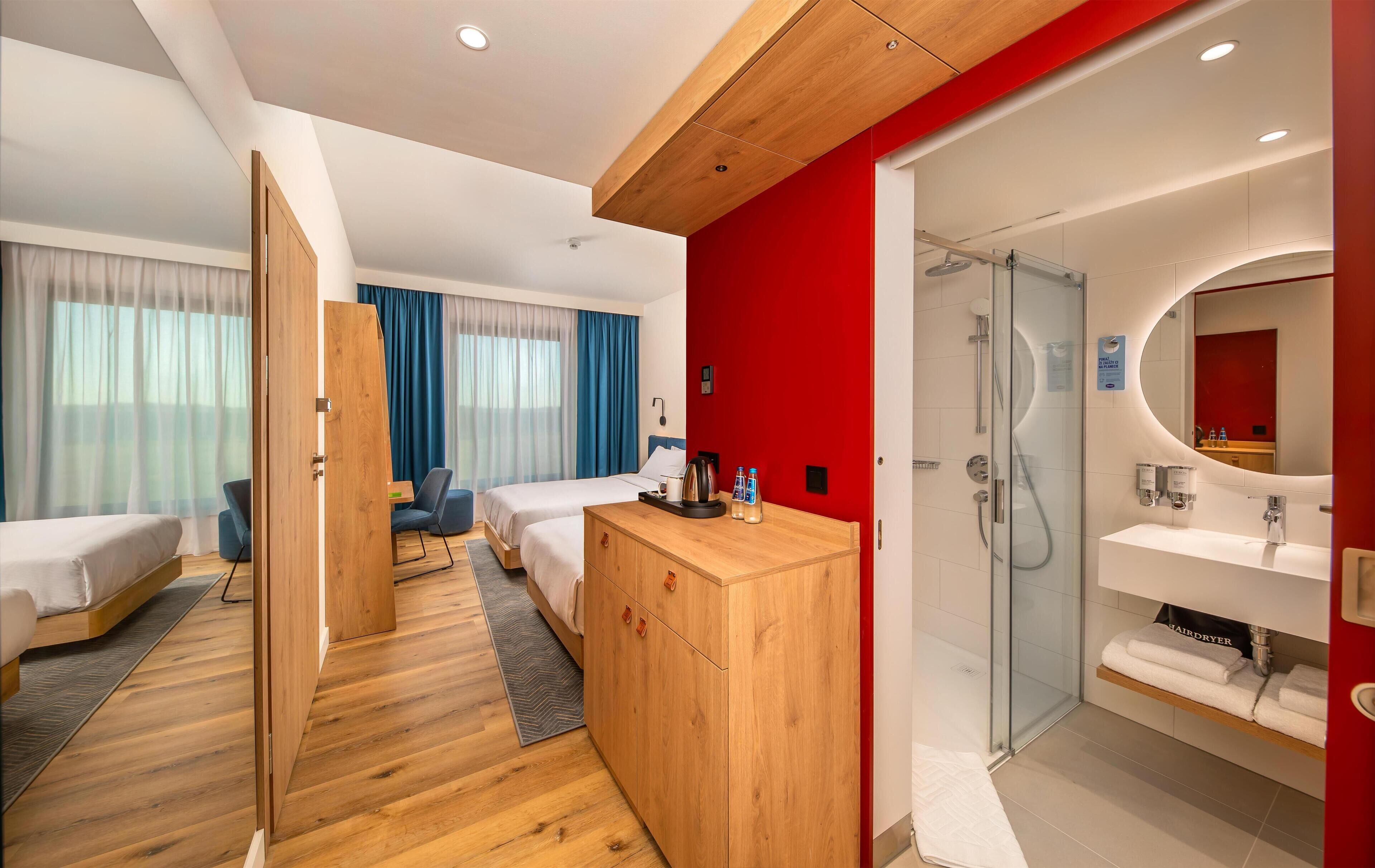 A hotel room with two beds, a wooden wardrobe, a desk, a chair, a shower area, and a bathroom with a mirror and sink.