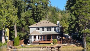 A two-story house with a dock extending into a lake, surrounded by dense trees.