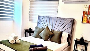 Un lit bien fait, avec une couvre-lit verte et des serviettes pliées, une tête de lit moderne et une petite table de chevet avec une lampe.