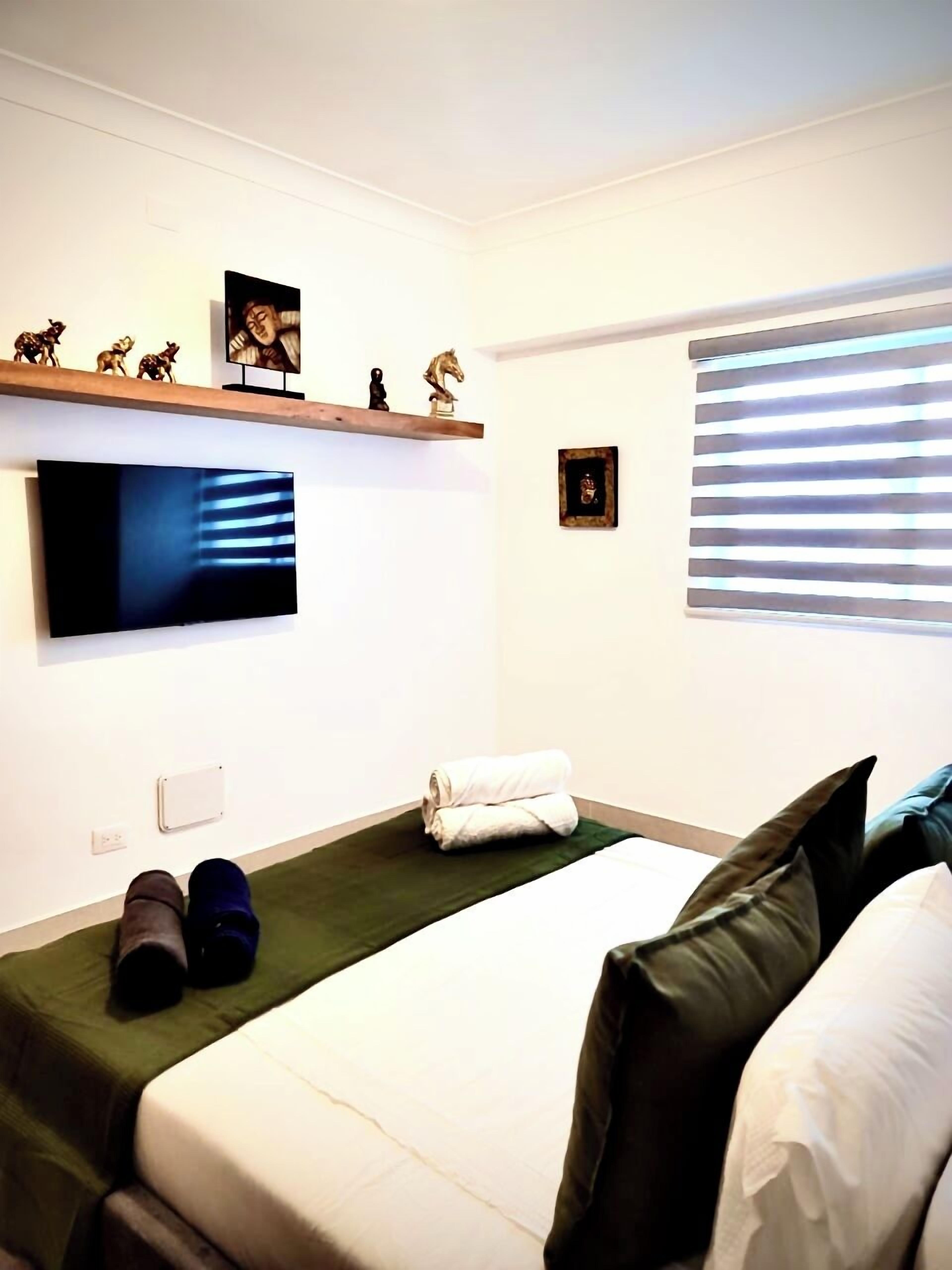 A hotel room with a bed, a TV mounted on the wall, a small shelf with decorative items, and a window with blinds.