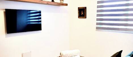 A hotel room with a bed, a TV mounted on the wall, a small shelf with decorative items, and a window with blinds.