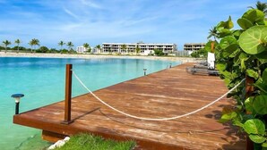 A wooden dock extending into a clear blue sea with a sandy beach and palm trees in the background.