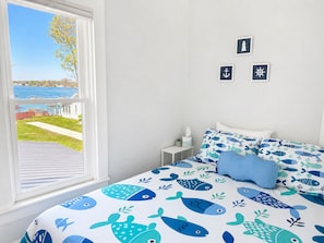 A bedroom with a bed featuring a blue and white nautical theme, a window with a view of a grassy area and a body of water, and three nautical-themed wall decorations.
