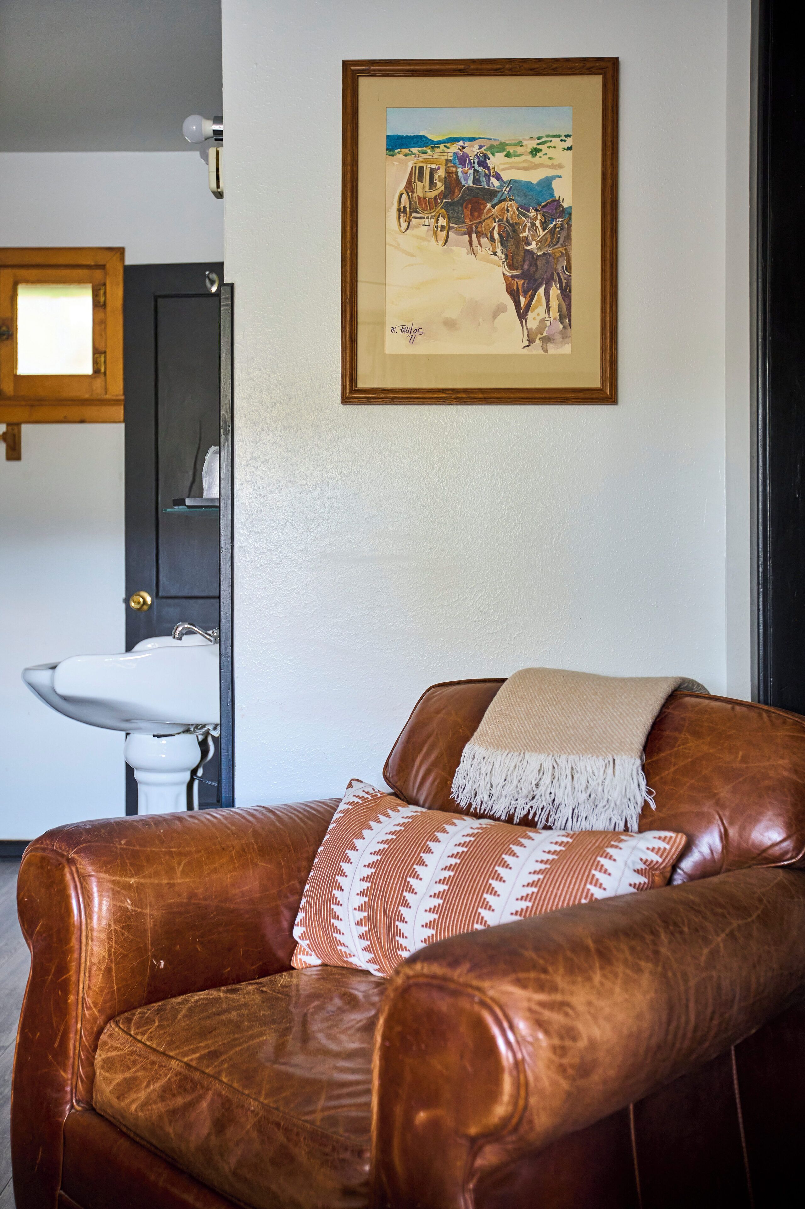 A brown leather armchair with a patterned throw pillow, a framed painting above it, and a white sink in the background.