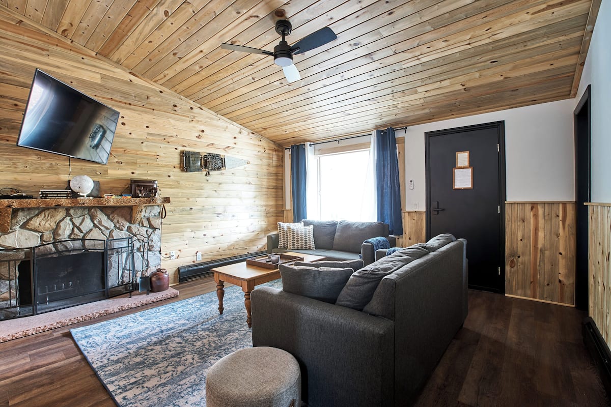 Un accogliente interno di una baita con camino in pietra, una moderna TV a schermo piatto, un divano grigio e un soffitto in legno.