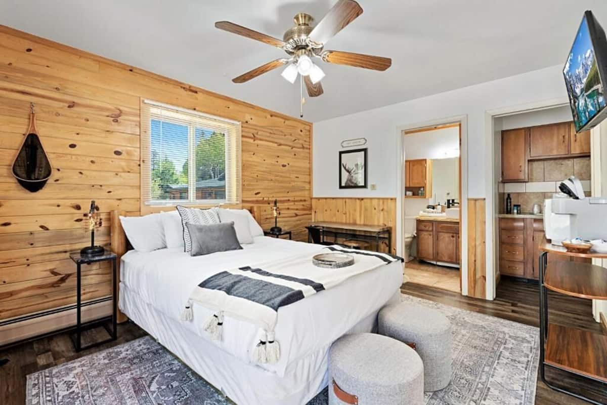 Una camera da letto con parete in legno, un letto, una ventola a soffitto, un comodino e una finestra con persiane.