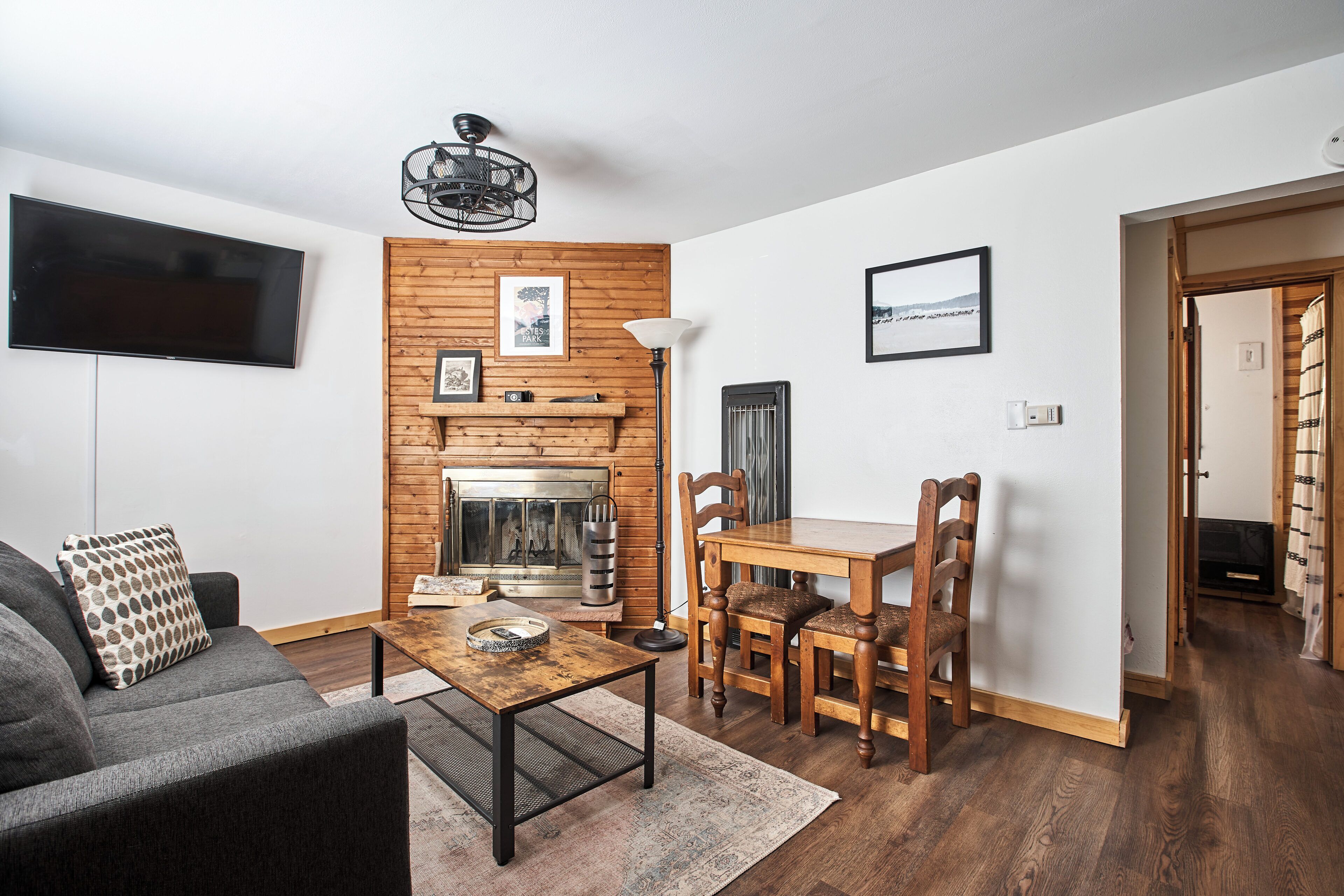 A modern living room with a flat-screen TV, wooden wall paneling, a fireplace, and a dining area with wooden chairs and a table.
