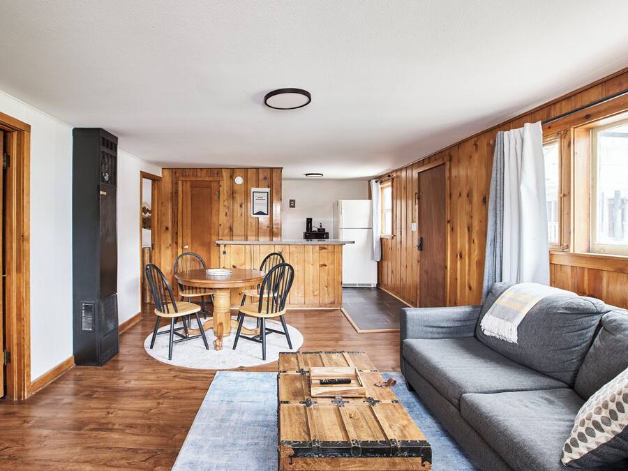 A cozy living room with a grey sofa, wooden dining table, and chairs. A small kitchen area is visible in the background.