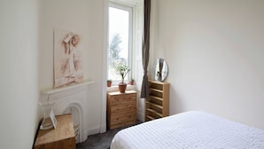 Une chambre à coucher avec un lit, une commode, un miroir, une fenêtre avec des rideaux et un tableau accroché au mur.