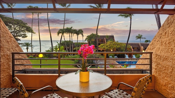 A balcony with a round table, chairs, and a vase of flowers. It overlooks a pool and palm trees, with a sunset view.