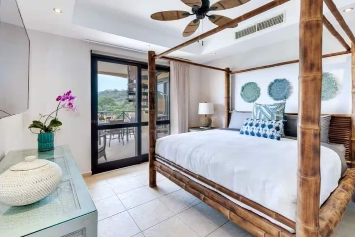 A four-poster bed with a wooden frame, a glass table with a white decorative item, a potted orchid, and a view of a balcony with outdoor furniture.