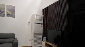 A living room with a grey sofa, a white air conditioner, a wooden cabinet with a TV, and a large window with blinds. - Luxury Solar Home where Style Meets Sustainability (Lipa)