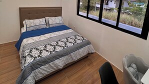 A modern bedroom with a large bed, a desk, and a window with a view of buildings and greenery. - Luxury Solar Home where Style Meets Sustainability (Lipa)