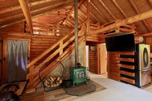 A wooden cabin interior with a staircase, a green wood stove, a black television, and a refrigerator.