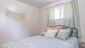 Un dormitorio con cama, almohadas, un colgador de macramé en la pared y una ventana con cortinas.
