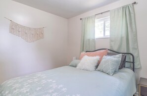Un dormitorio con cama, almohadas, un colgador de macramé en la pared y una ventana con cortinas.
