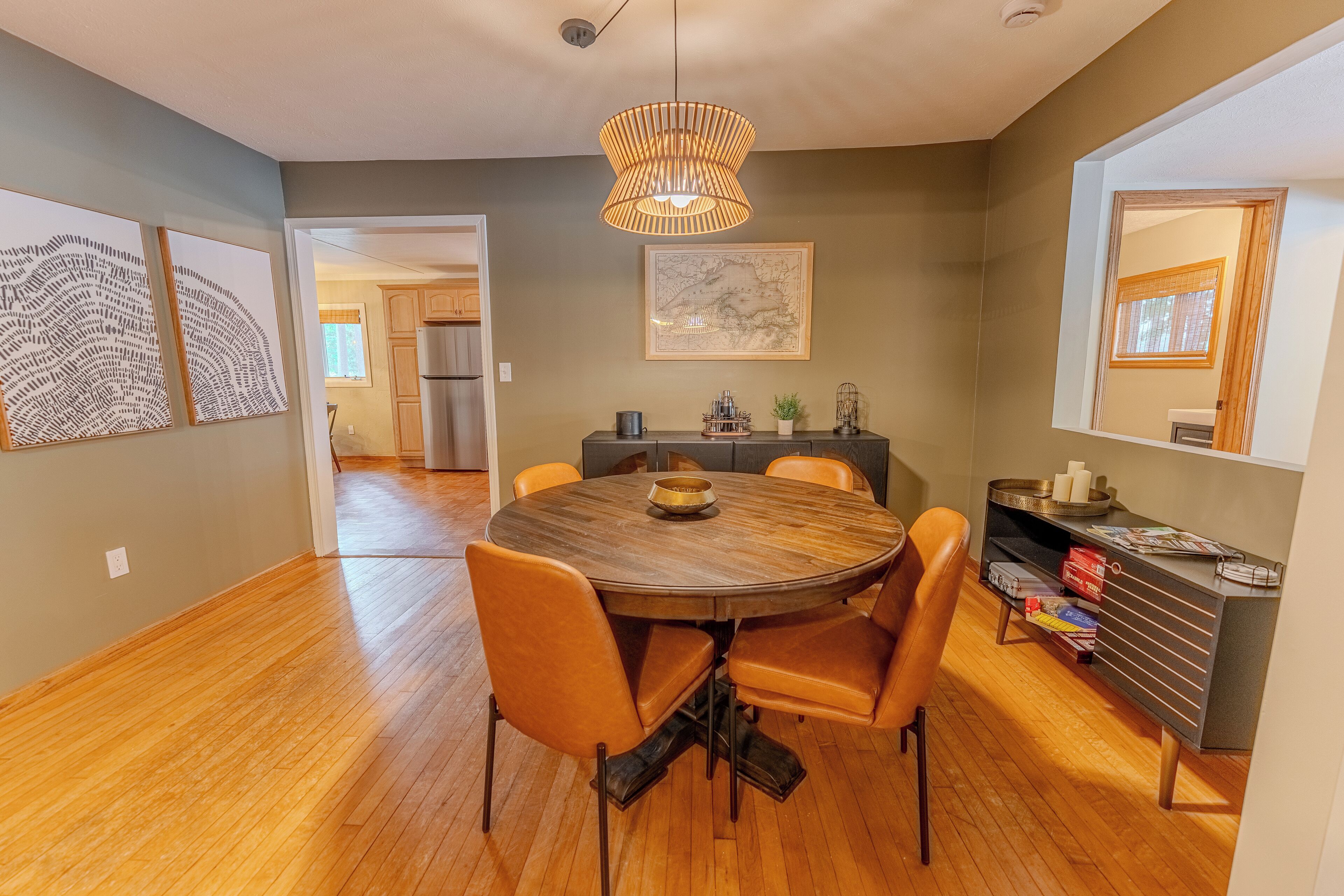 A dining area with a round wooden table, four orange chairs, a hanging light fixture, and a bookshelf with books and decorative items.