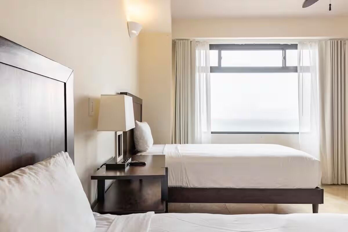 Habitación de hotel con dos camas, una mesita de noche con lámpara y una ventana con cortinas.