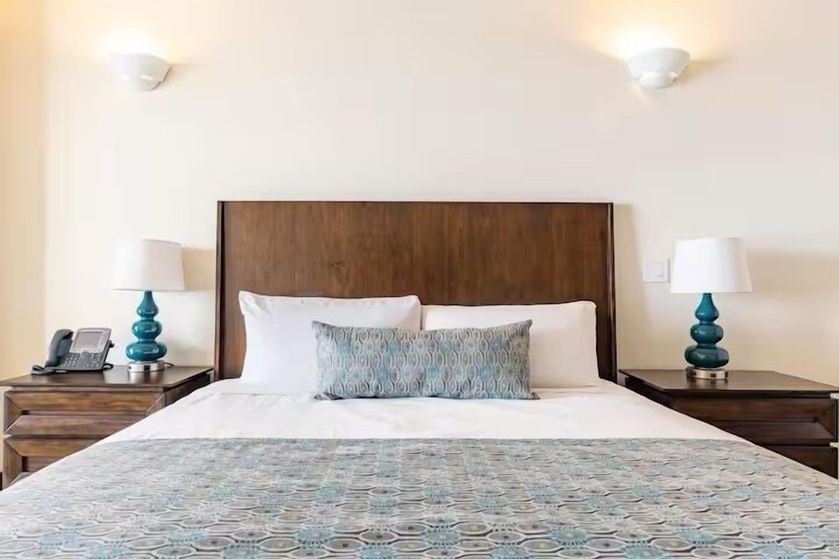 A hotel room with a wooden headboard, a patterned bedspread, two bedside tables with lamps, and a telephone on one of the tables.