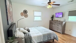 Un dormitorio con una cama, un aparador, un ventilador de techo, un televisor y una ventana.