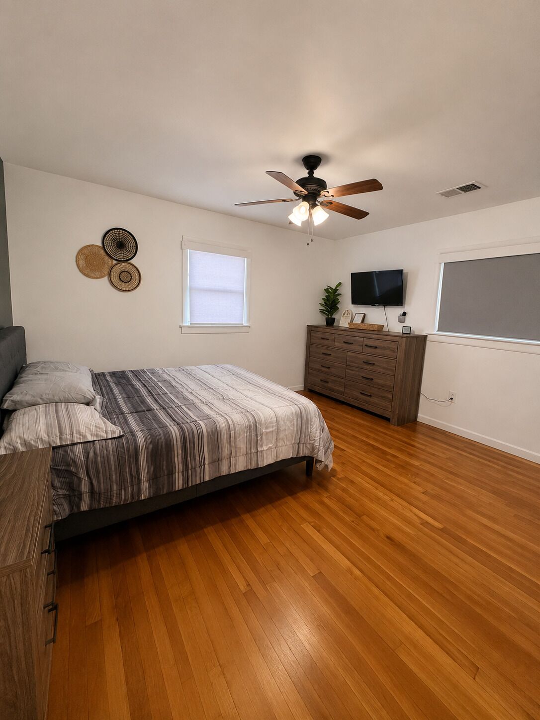 A bedroom with a bed, wooden flooring, a ceiling fan, a dresser, a television, and wall decor.