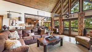 Ein geräumiges Wohnzimmer mit einem großen braunen Ledersofa, einem Holztisch und Blick ins Freie durch große Fenster.