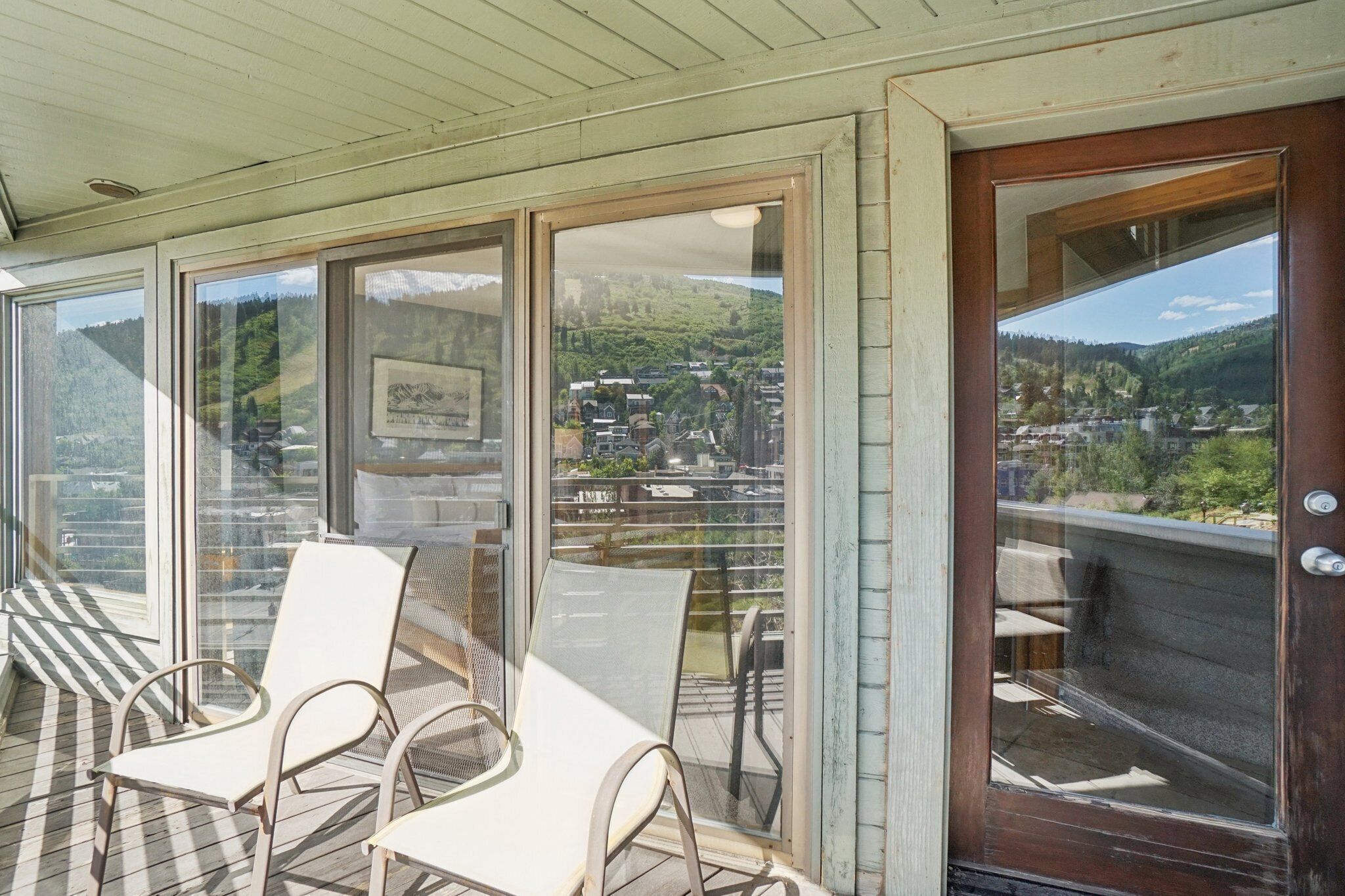 Un área de estar al aire libre con techo, sillas y una mesa, grandes ventanales con vistas a un pueblo y colinas, y una puerta de madera.