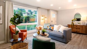 Una sala moderna con sofá, sillón y mesa de centro, un ventanal con vista a follaje, y un aparador de madera con objetos decorativos.