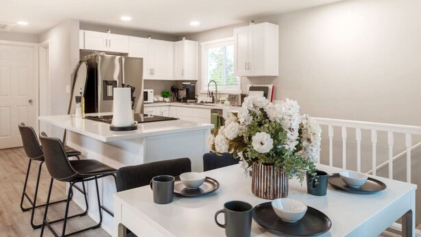 A modern kitchen with white cabinetry, a central island, and a dining area with a table set for two. - Modern 5BR 3BA Remodeled Home with BBQ Backyard (Seattle)
