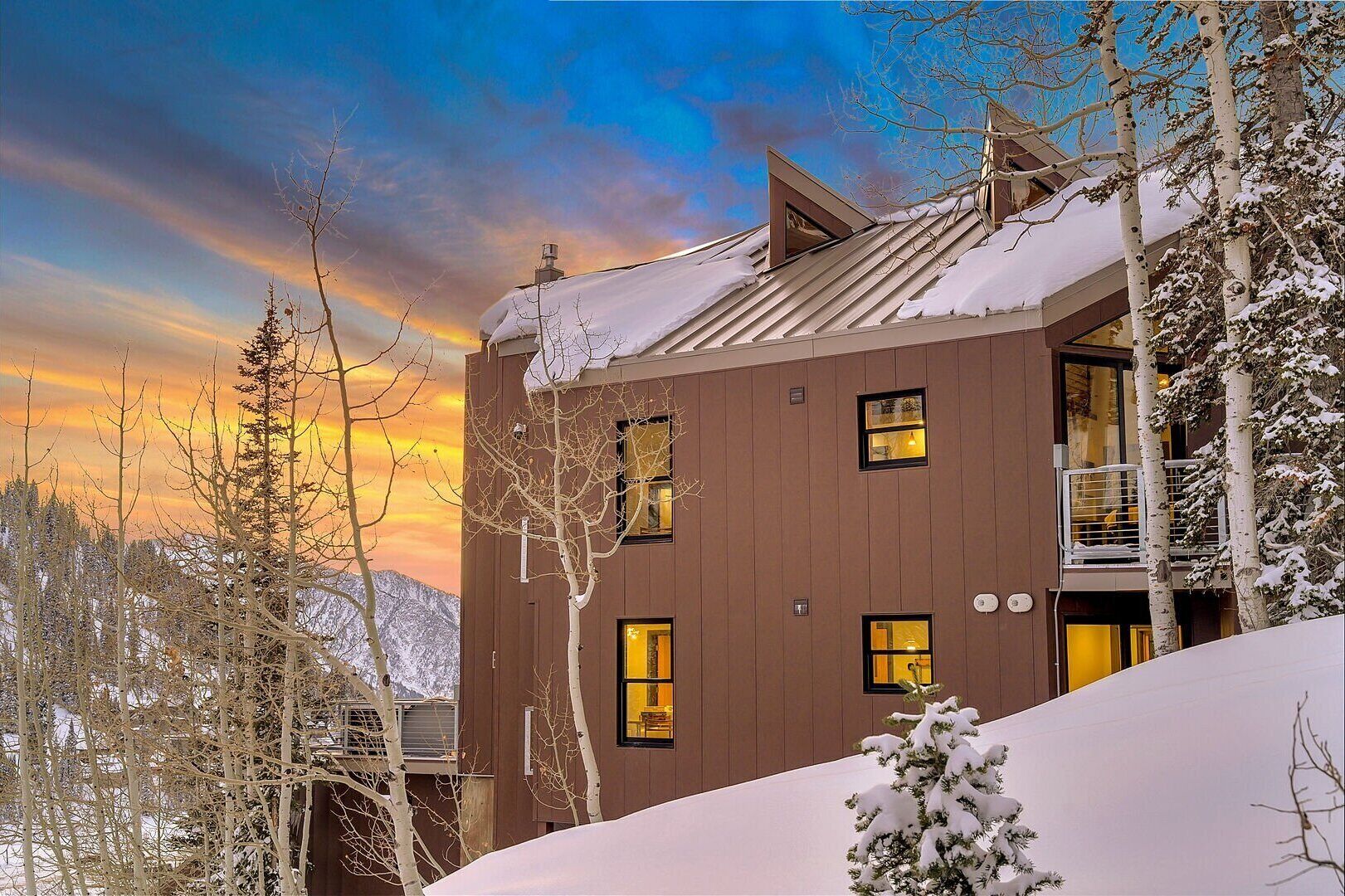 A modern wooden building with multiple windows and a balcony, surrounded by snow-covered trees and a mountainous backdrop.