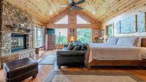 A log cabin interior with a stone fireplace, a bed, a leather sofa, and a ceiling fan. - STERLING LODGE - LUXURY MOUNTAIN VIEW CABIN (Sautee Nacoochee)