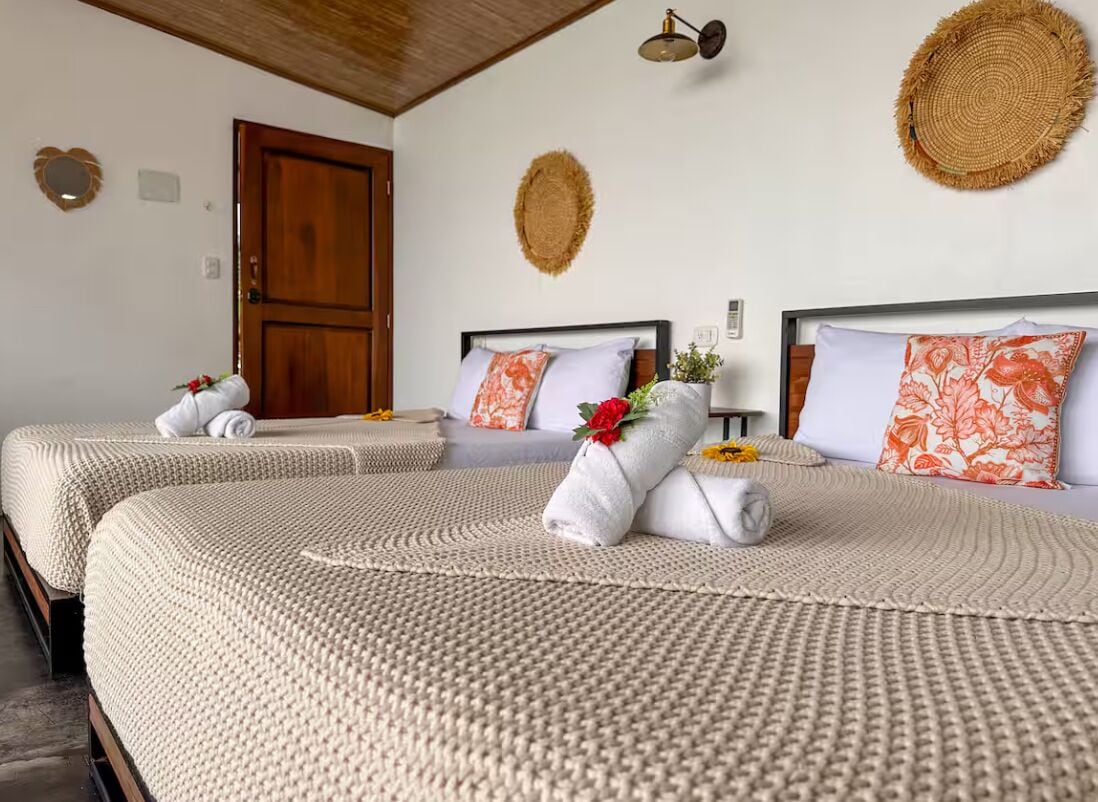 Una camera d'albergo con due letti, ognuno con una coperta beige a texture e un asciugamano bianco arrotolato con un fiore rosso.