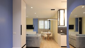 A modern living room with a grey sofa, a small dining table with chairs, and a kitchen area with a microwave and a flat-screen TV. - Illyrian Apartment 02 (Durrës)