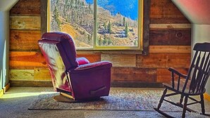 A log cabin room with a large window offering a mountain view, a purple chair, and a wooden rocking chair. - Anvil Room Rustic Stay at Columbine Lodge (Silverton)