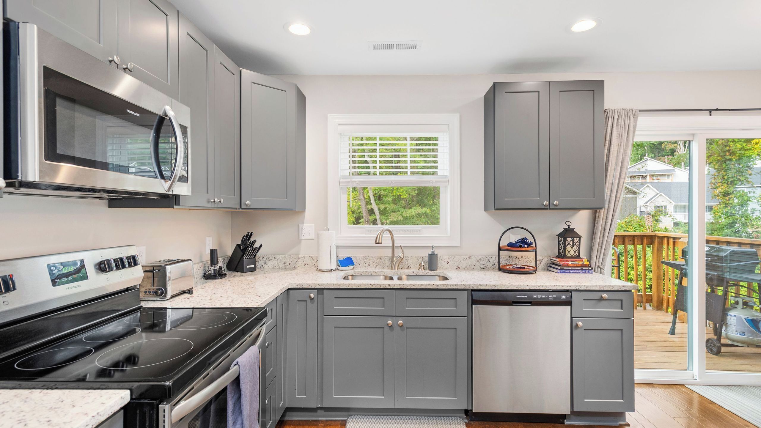 A modern kitchen with grey cabinets, a stainless steel microwave, a built-in oven, a dishwasher, and a window with blinds.