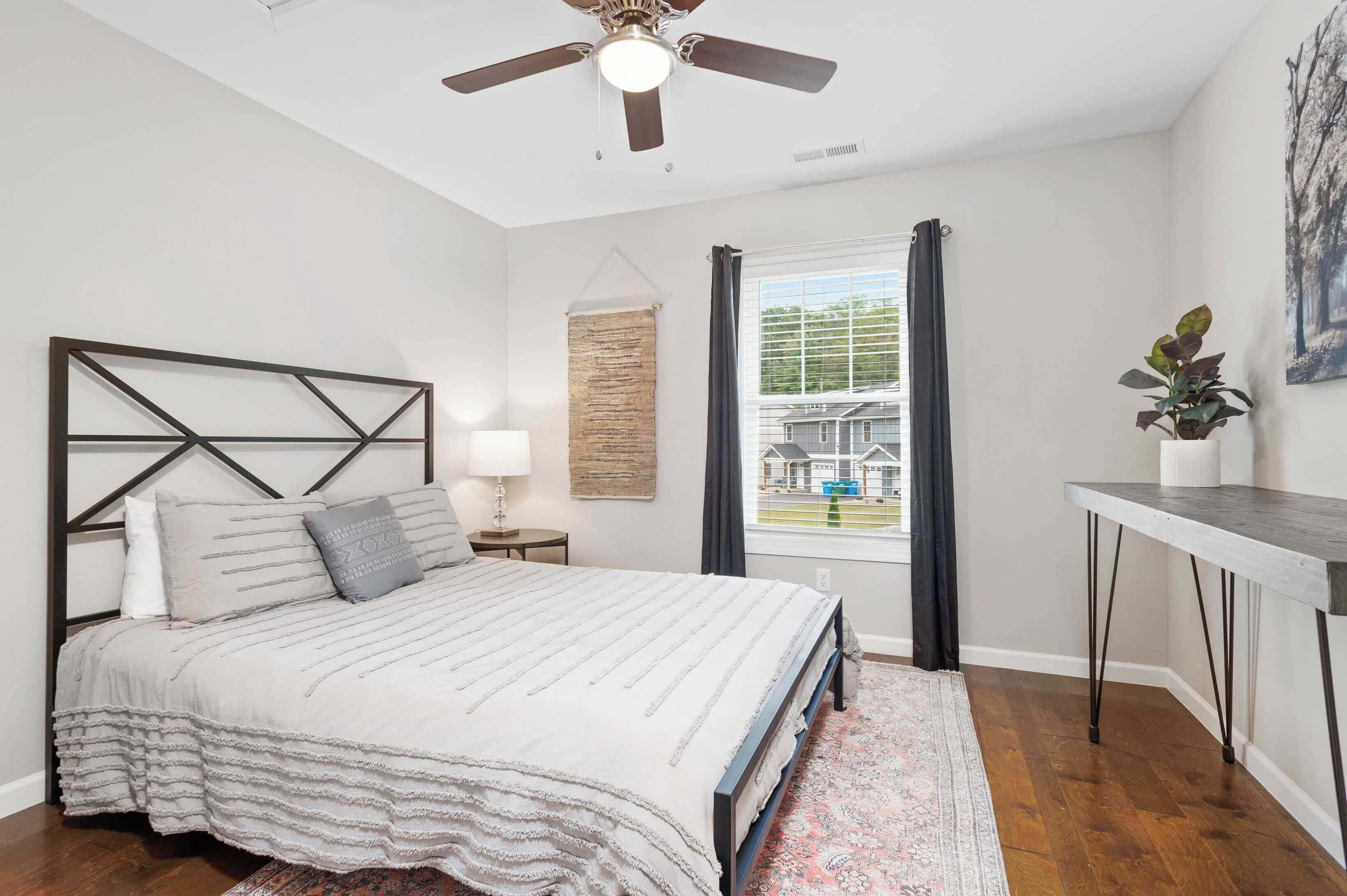 A bedroom with a bed, a ceiling fan, a window with curtains, a bedside table, a lamp, a plant, and a painting on the wall.