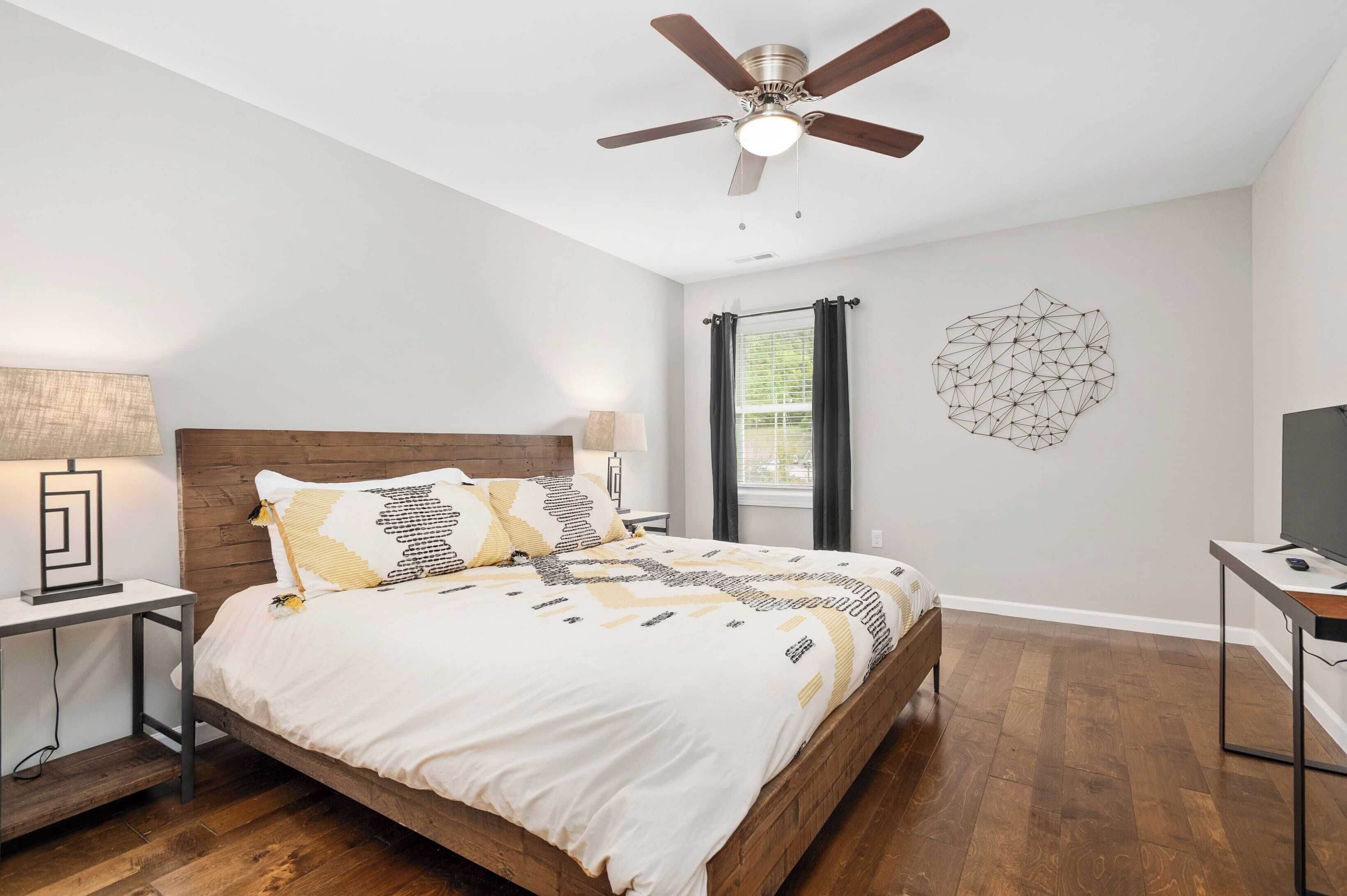 A bedroom with a wooden bed, a ceiling fan, a wall art piece, and a window with curtains.