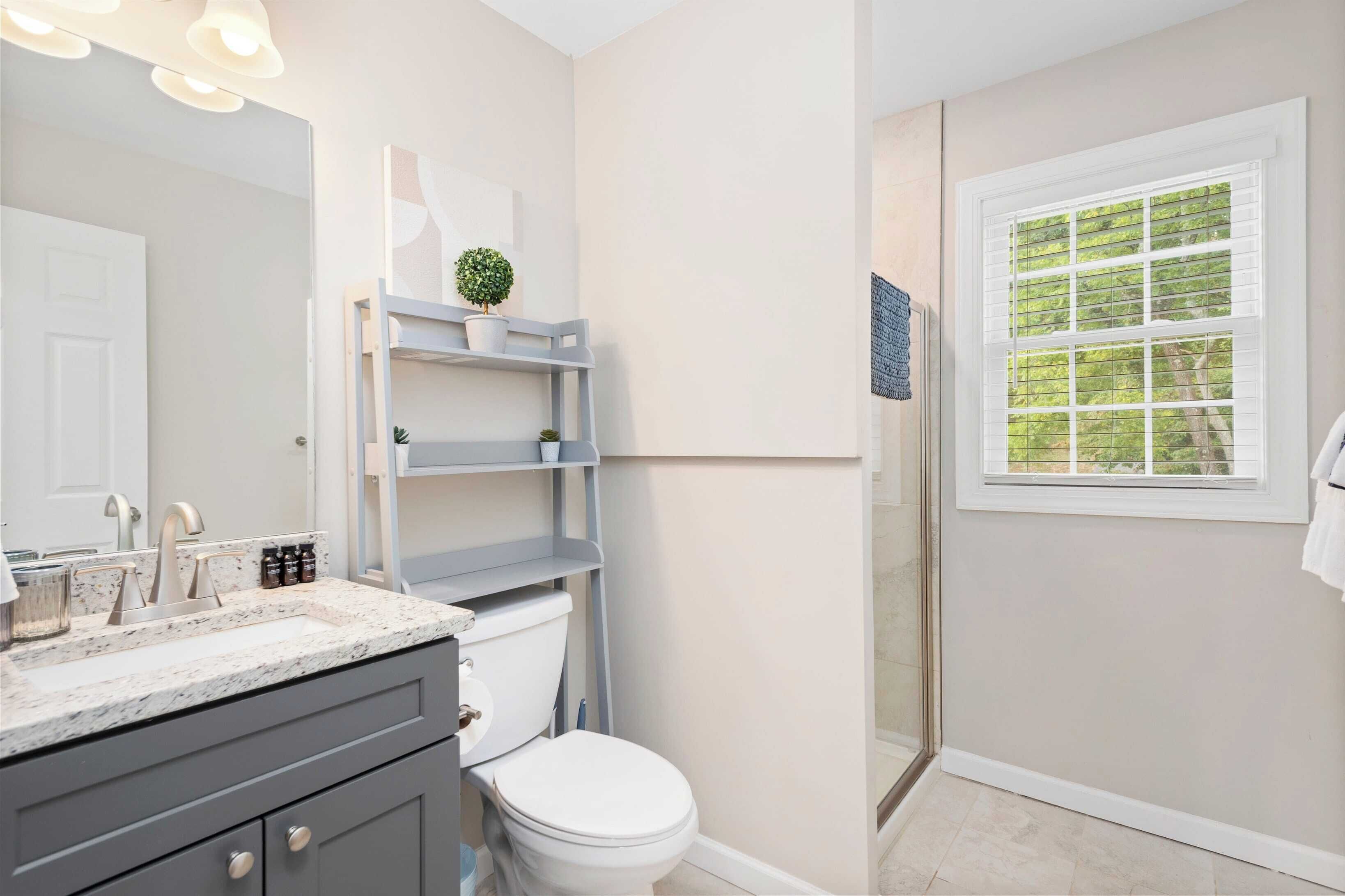 Une salle de bain avec des toilettes blanches, un lavabo doté d’un plan de travail en marbre, un miroir et une fenêtre avec des stores.